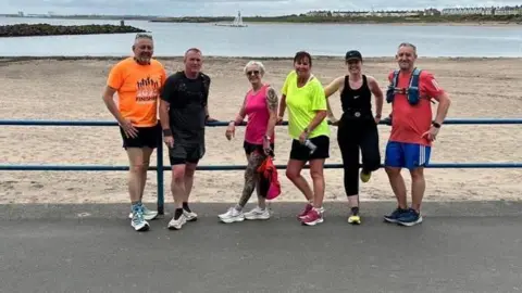 Karen McNeill Smith  A group of six runners wearing bright coloured clothes standing in front of the beach at Newbiggin-by-the-Sea in Northumberland.