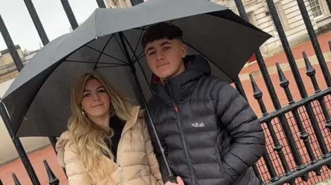 Devon Andrews Cherish and Ethan stand under an umbrella. 