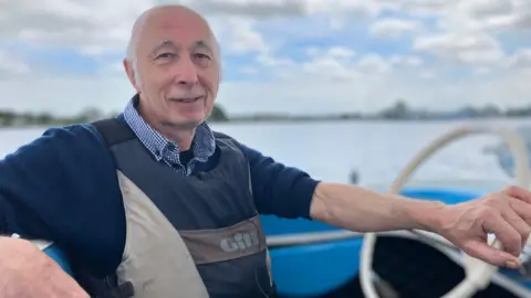 A man with grey hair is sat in the driver seat of a blue and white boat with his hand on the steering wheel. He is wearing a blue and white checkered shirt with a navy-blue jumper and a life jacket on top. 