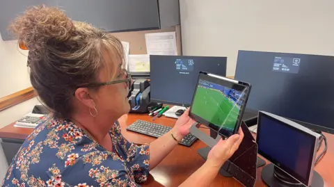 Heather Earhart Heather Earhart, 54, wears a floral shirt and glasses, her curly blonde hair tied up, as she sits at her desk holding a tablet that's showing the England versus Argentina match