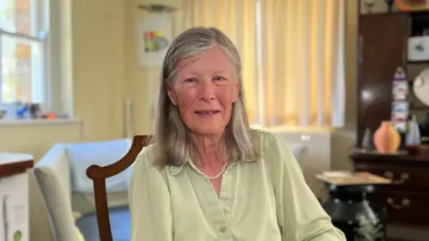 Mary Venturini sits in a wooden chair in a bright living room. She has shoulder-length grey hair, is wearing a light green button-up blouse, light blue jeans, a necklace and a watch.