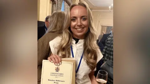 A young woman with long blonde hair holds a certificate. She is wearing a white shirt and has white painted fingernails. She is standing a room of people and has a broad smile on her face.