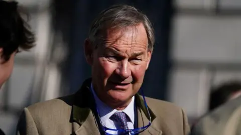 PA Rupert Lowe wears a beige jacket and a checked shirt and tie. His spectacles hang around his neck.