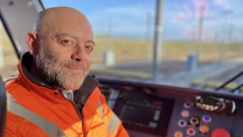 Tram driver Michele Mastromarino sitting in the cab of his tram. He is bald and has a short grey beard and moustache. He is wearing a high-vis orange jacket over a white shirt and grey tie