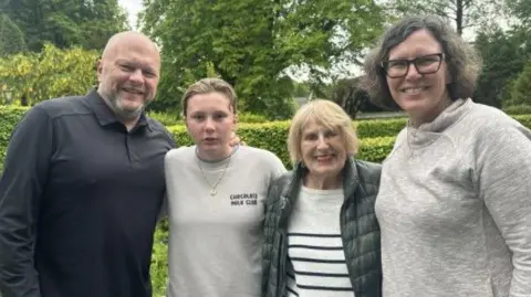 Four people stand together in front of a green hedge. Jason Camis and his wife Molly are at either end, with Luciana, their daughter next to Jason. Next to Molly is Joan Carus, an older woman with blonde hair and red lipstick wearing a black jacket and a white striped jumper They're all smiling except Luciana, who looks surprised.