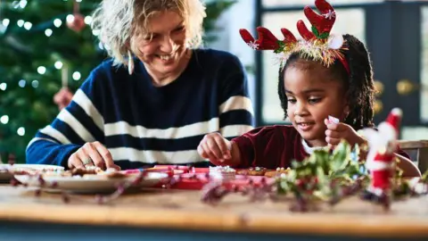Getty Images A grandmother helps her granddaughter to decorate Christmas cookies. The girl is wearing reindeer antlers and is picking up decorations. There is a Santa model on the table and a tree behind them.