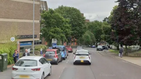 Rouen Road, Norwich, with several cars on the road, people walking up and down the street, trees are along the road, and a large brick building to the right.