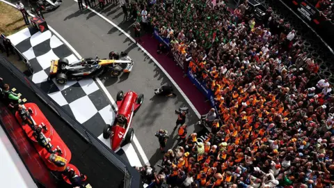 Getty Images A shot from above of the podium at the British Grand Prix. A Formula One car is visible alongside a classic red F1 car. A large group of people are to one side looking towards the podium.
