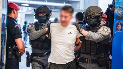 Central Investigative Bureau, Royal Thai Police She Zhijiang, whose face has been blurred out, wearing a white Adidas t-shirt, escorted by two Thai officers in combat gear and face masks.