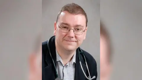 Hull University Teaching Hospitals NHS Trust A portrait photo of a man with short brown hair and rectangular glasses smiling into the camera. He has a medical device around his neck and is wearing a check shirt and a black jacket.