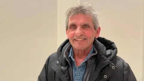 Denny Wilson smiling at the camera. He has short grey hair and blue eyes ans is wearing a black puffer coat and a denim shirt.