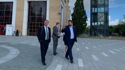 Simon Dedman/BBC A group of four people in the centre of the picture. Two in front are starting to walk and are looking around a square. they are men in suits.