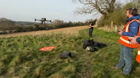 A student in a field wearing an orange hi-vis vest flies a drone a short distance in front of him.