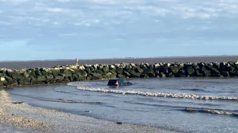 Lara King/BBC A black car can be seen submerged by waves next to a sea wall of rocks. The beach can be seen in the foreground. 