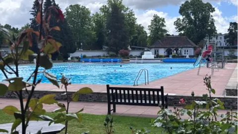 Pink roses are visible in the foreground with a bench, pool and lifeguard in the background.