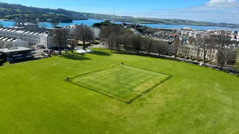 Plymouth Cricket Club Plymouth Cricket Club's field is luscious and green with the wicket cordoned off. The sea is visible in the background as well as lots of buildings and trees.