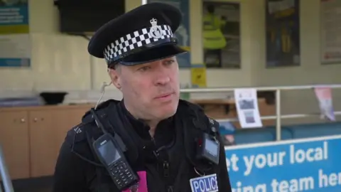 PC Ben Chadwick of Devon and Cornwall Police is looking away from the camera. He is wearing the usual police uniform with a cap and radio equipment. He is stood in front of a police information stand with promotional posters in on it.