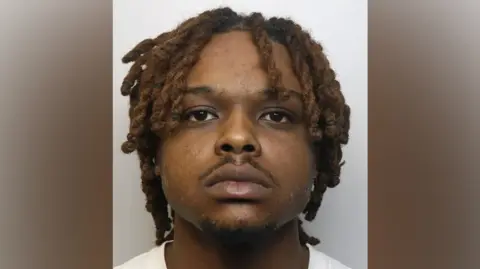 A police custody photograph showing a black man with dreadlocks and a small moustache, staring blankly forward 