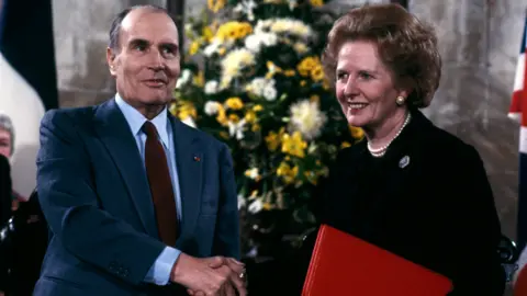 Margaret Thatcher and the late French President, Francois Mitterrand, shake hands. Thatcher is carrying a red file in her other hand.