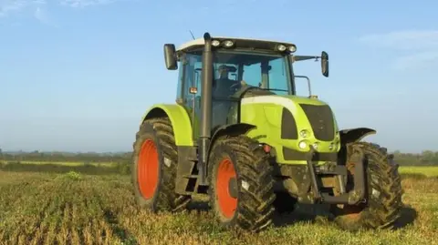 A green tractor with red wheel hubs in a field with a worker at it's wheel. Crops can be seen growing in lines. The sky is blue with light clouds.