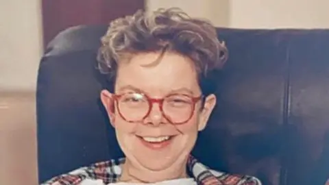 A photo from the 80s of a woman in a leather arm chair wearing round red glasses and smiling