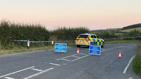 Police car stationary on road which has been cordoned off with blue and white police tape. Blue signs have been put up on the road saying Police Accident and Police Road Closed in English and in Welsh alongside orange and white cones. There is a bend in the road and hills and fields are visible behind.