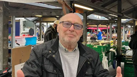 Elizabeth Baines / BBC A man in a grey top stands in front of a stall. He wears a green coat.