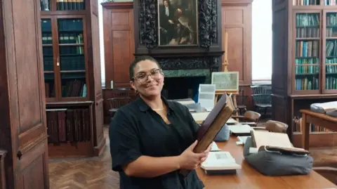 Heather Marks is in the Bristol room of the Central Library. She has short hair and large tortoise shell glasses. She is wearing a navy blue shirt and holding a large book with a burgundy cover. She is standing next to a large table and there is a painting on the wall in the background.