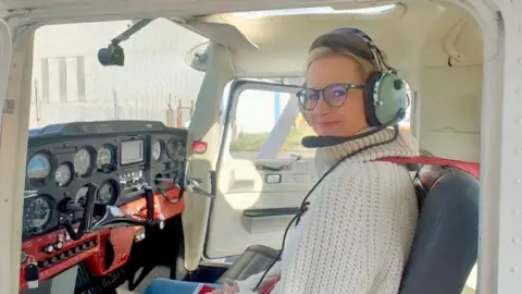 Christina Mackaill Christina Mackaill in a simulation aircraft. She is wearing glasses, a cream wool jumper and headset. 