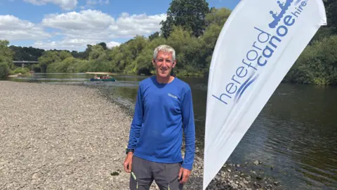 A man with grey hair is standing by a white banner, including the words Hereford canoe hire on the right. He is wearing a blue top and grey trousers and standing near a river.