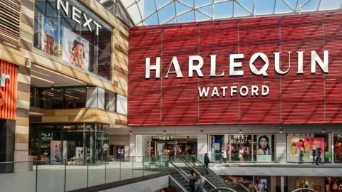 Red internal wall of the Atria centre, with lettering saying "Harlequin Watford". The Q in Harlequin has a diamond-shaped "hole" in the middle, reflecting the traditional Harlequin checked pattern. There is a next store below the sign, featuring pictures of models wearing Next clothing. There is an escalator in the foreground and another shop to the left of the picture.