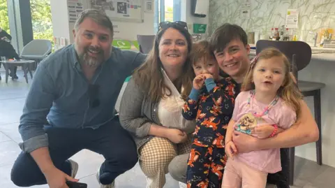 REBEKAH DALRYMPLE The family, Andy a man with grey hair and a beard, Rebekah with brown hair, Hector with short brown hair, Hugo a young adult with short brown hair, and Dotty a young girl with light hair and a pink t-shirt, they all smile brightly.