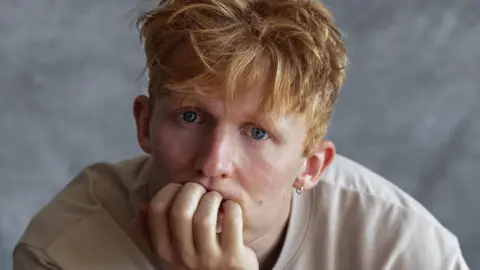 Adam Tucker looks thoughtful. He is resting his chin on his hand. He has blue eyes and gigner hair.