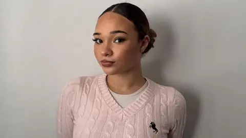 Demi, 17 is smiling at the camera, wearing a pink v-neck sweater and has her hair in a ballet bun.