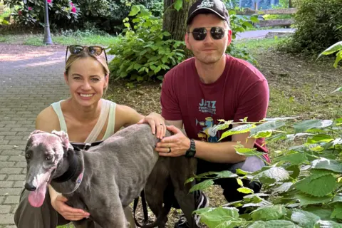 Supplied Natalie Angst and Tomas Antonic crouch beside Kiki the greyhound on a paved path in a leafy park. Kiki, wearing a collar, stands in the foreground, while the couple hold and steady her. Trees, dense greenery, a lamppost, and a wooden sign are visible in the background. Natalie Angst is smiling and wearing sunglasses on her head and a yellow top. Tomas Antonic has sunglasses on and a brown cap, black watch and maroon t-shirt.