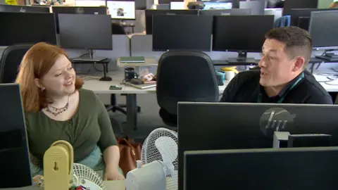 Two people in an office sit facing each other in an open-plan office, talking across computer screens. 