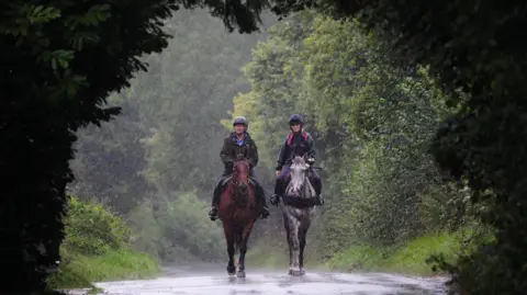 PA Media Two female horse riders are seen walking their horses towards the camera. It is raining heavily and the riders are framed by dark foilage