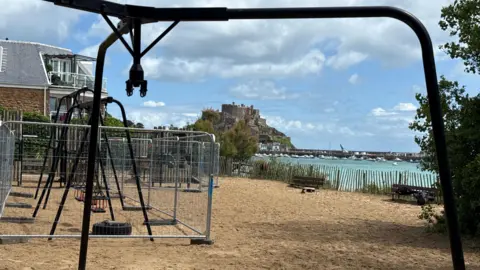 A play park on a Jersey seafront. The play equipment is fenced off. A castle can be seen in the distance. The floor of the park is covered with sand.
