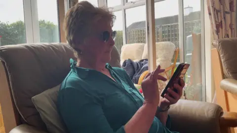 An elderly woman called Ruth Goodman sits on a chair in her living room while looking at her mobile phone. She has glasses on and is wearing a teal blouse.