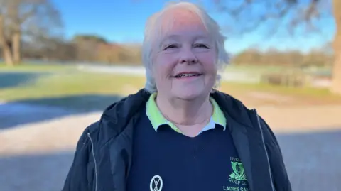 GEORGE CARDEN/BBC Gillean Fuller, a woman with short grey hair, wears a blue overcoat over a blue jumper and green golf shirt, and looks directly into the camera.