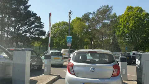 BBC A driver's view of a queue of traffic entering a car park. Cars are waiting at open entrance barriers unable to move because of a queue of cars snaking around the outdoor surface car park.