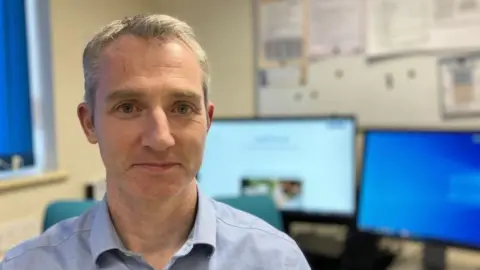 Dr Campbell Murdoch sitting in his GP practice wearing a blue shirt. Behind him is a computer screen. He is faintly smiling at the camera.