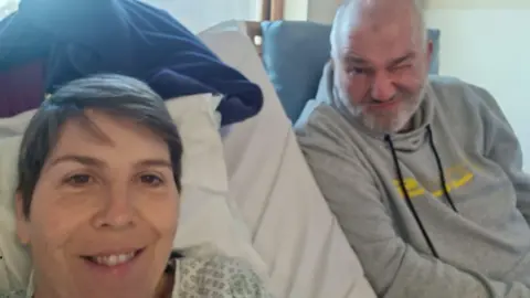 Family photo Serena sits in a hospital gown in her hospital bed with her husband smiling and winking next to her.