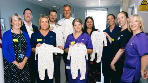 AFC Bournemouth A group of hospital staff and people from AFC Bournemouth stand together in a ward for a posed picture. Two women at the front of the group are displaying AFC Bournemouth sleepsuits. 
