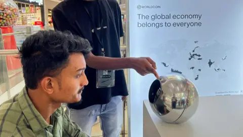 A man in India sitting before an earlier World device, a silver orb, to have his eye scanned as a form of human identification. He has dark hair and a green shirt, and looks straight into the orb, on a desk in front of him, while another man points to it. 