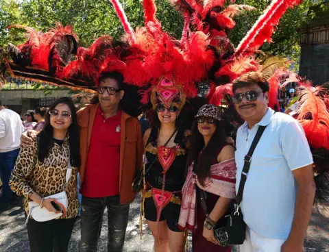 A group of parade participants pose for a photograph beside a performer wearing an elaborate red feathered headdress and costume. The large red and black feathers fill much of the background, with trees and festival crowds visible behind.