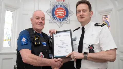Essex Police Allan shakes hands with Chief Constable Ben-Julian Harrington as they both hold a framed certificate from the National Police Staff Awards 2025. They are in a white room with the Essex Police emblem on the wall. It is blue, white and red. Allan wears his uniform minus his hat, and Mr Harrington wears a short sleeved white police shirt with a black tie. He has short brown hair, is clean shaven, and wears a grey smart watch.