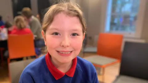 A young girl with light brown hair tied back from her face is smiling at the camera. She is wearing a blue sweatshirt with a red t-shirt underneath.