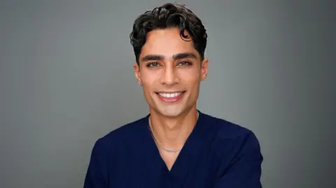 Zayn Khalid Majeed Dr. Zayn sonríe a la cámara. Lleva un inserto quirúrgico azul y una cadena de plata alrededor de su cuello. Tiene dientes blancos, piel oliva y un ligero rizo en su cabello castaño. También tiene un pequeño aro plateado en la oreja.
