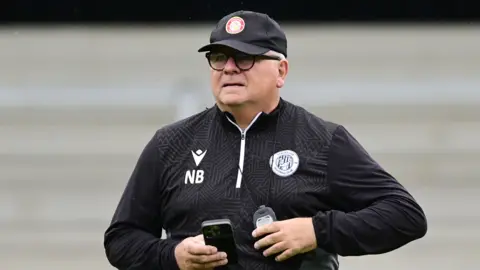 Neil Banfield wearing a black top with a Stevenage badge and NB written on it and a black cap. He is holding a phone in his hand